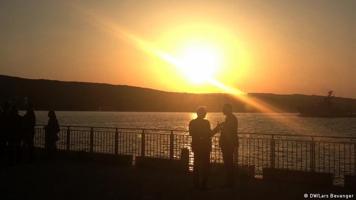 At a very low angle, the bright orange sun backlights a distant peninsula and a pair of men talking on a seaside boardwalk.
(Photo: DW / Lars Bevanger)
