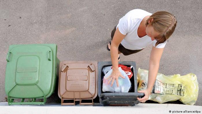 Eine Frau legt eine Tüte Müll in eine Mülltonne. Daneben stehen verschiedene andere Mülltonnen. 