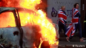 Women draped in Union flags walk past a burning car after loyalist protesters attacked the police with bricks and bottles as they waited for a republican parade to make its way through Belfast City Centre August 9, 2013 (Photo: REUTERS/Cathal McNaughton)