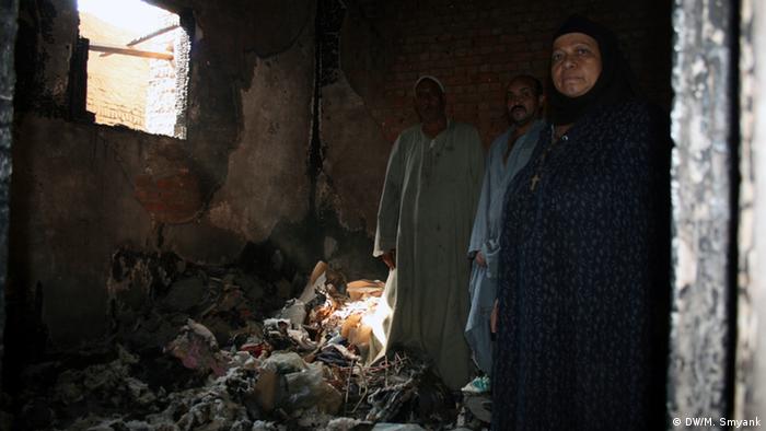 A Middle Eastern family wearing robes stands in the darkened and rubble-filled room of their destroyed former house.
(Photo: Markus Symank / DW)