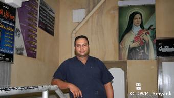 Standing next to a picture of the Virgin Mary affixed to the wall, a man of Middle Eastern descent wearing a blue, short-sleeved collared shirt rests his arm on a metal rail in a beige room.
(Photo: Markus Symank / DW)
