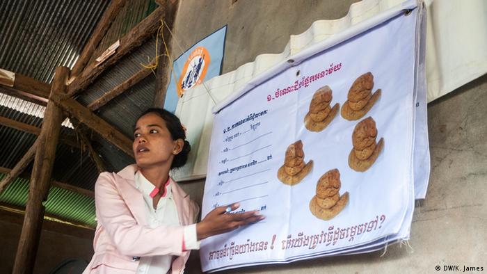 A cambodian women stands next to a poster with pictures of feces, giving a presentation to villagers about health and sanitation (photo: Kyle James)