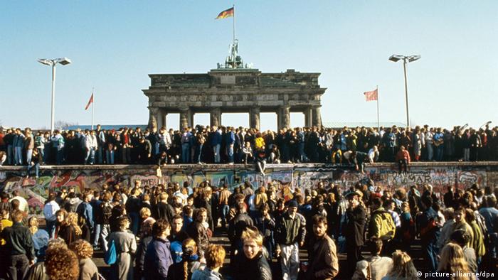 Thousands celebrate the falling of the Berlin Wall in a color photograph with the Brandenburg Gate in the background.