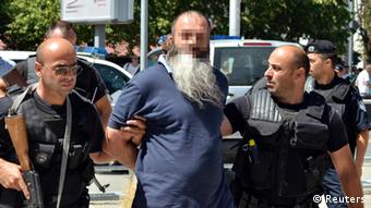 A bearded middle-aged man of Middle Eastern descent is led away, handcuffed, by police